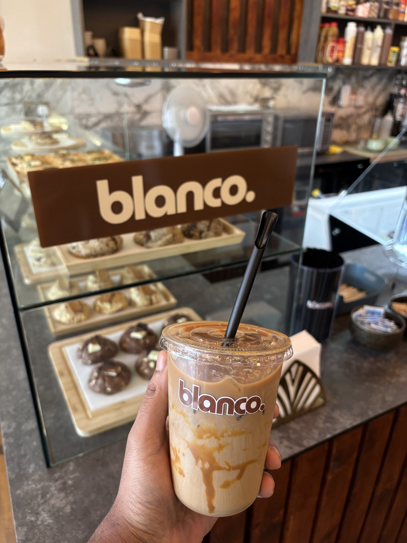An iced Blanco latte held in front of the pastry case and the house blanco. sign.