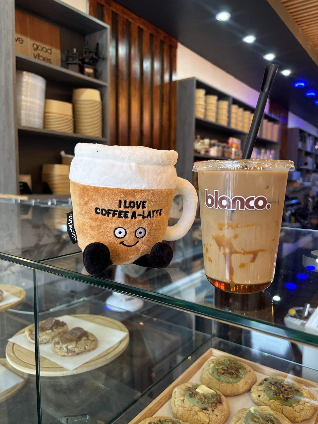 The Blanco mug mascot beside an iced latte, with cookies in the case below.